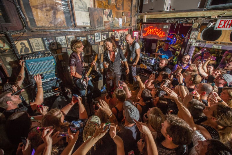 A band performs energetically in a small, crowded venue with enthusiastic fans reaching out and taking photos. The walls are adorned with posters and photos. Colorful lights enhance the lively atmosphere.