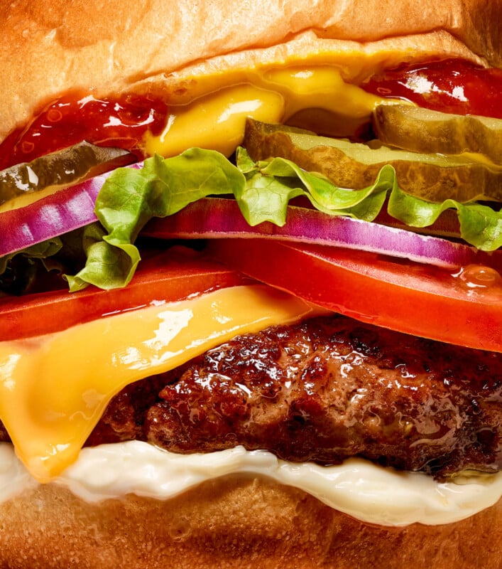 Close-up of a juicy cheeseburger with melted cheese, lettuce, tomato, pickles, red onion, ketchup, and mayo in a bun. The vibrant colors and textures highlight the freshness of the ingredients.