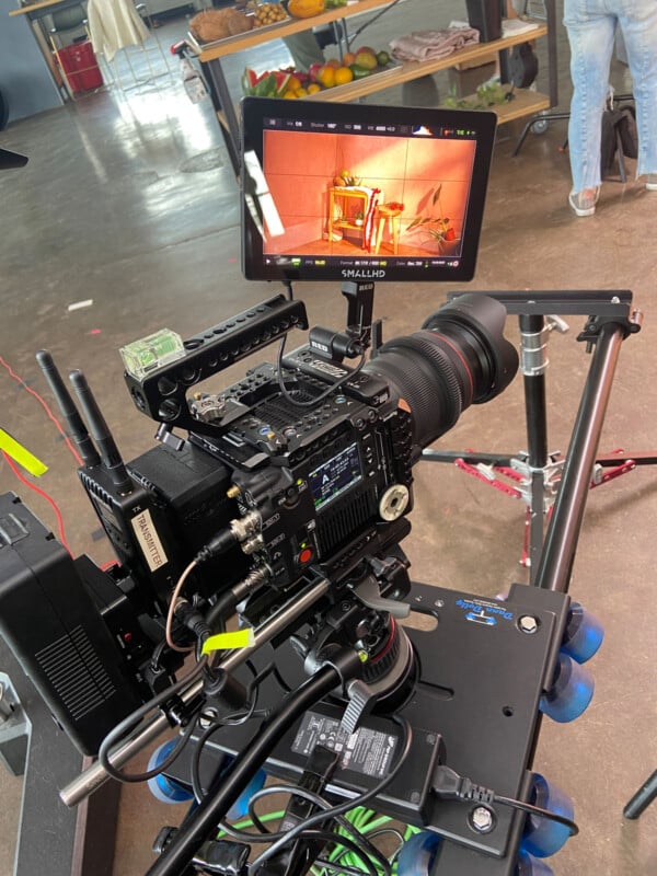 A professional camera mounted on a dolly in a studio setting, focused on a small monitor displaying a live feed of a chair in front of a pink backdrop. Various cables and filming equipment surround the setup.