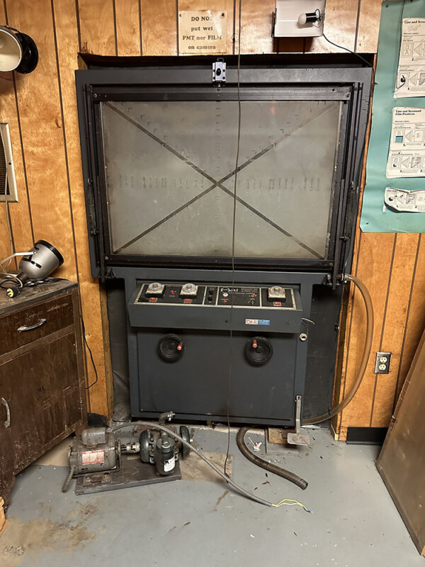 Old wooden room with a large, dusty industrial machine. It has a control panel with buttons and dials, situated beneath a retractable screen. Nearby, a small motor or pump on the floor, with cables and hoses attached, adds to the vintage setup.