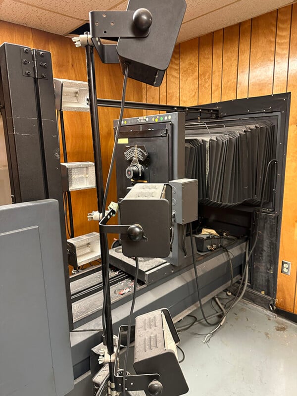 A vintage X-ray machine with a large vertical frame and multiple adjustable lights is set up in a room with wood-paneled walls and a gray floor. The machine's various components are visible, including accordion-style sections.