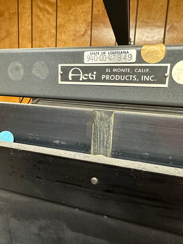 Close-up of a vintage piece of equipment from Acti Products, Inc., located in El Monte, California. It has a label from the State of Louisiana with the number 940004949. The background features wood-paneled walls.