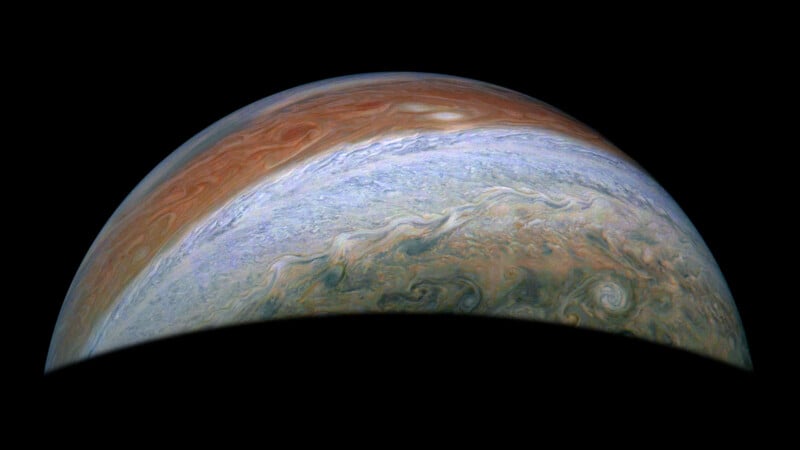 A high-resolution image of Jupiter showcasing its swirling clouds and vivid colors. The upper half displays bands of red, brown, and white, with complex swirling patterns throughout. The bottom is in shadow, highlighting the planet's curvature.