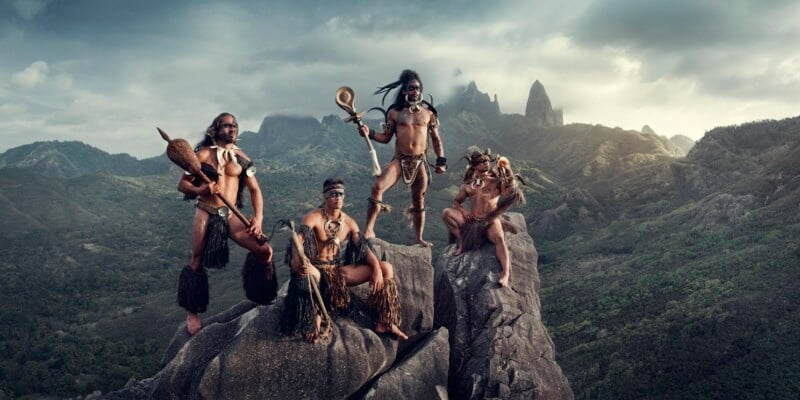 Four individuals in traditional tribal attire and holding various items pose on rocky cliffs with a lush mountainous landscape in the background. The sky is cloudy, creating a dramatic atmosphere.