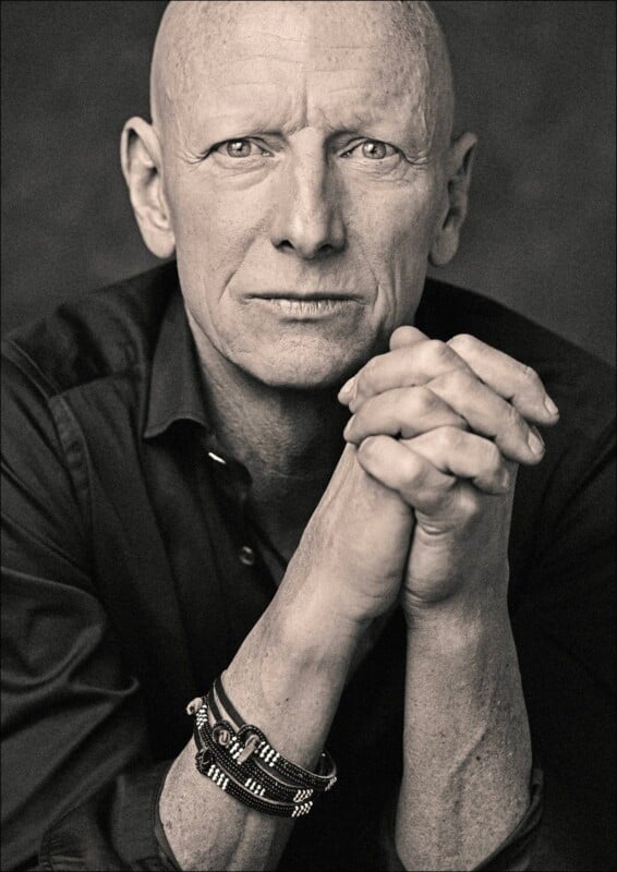 Black and white photo of a bald man with a serious expression, resting his chin on his clasped hands. He wears a dark shirt and multiple bracelets on his wrist. The background is a simple, dark tone.