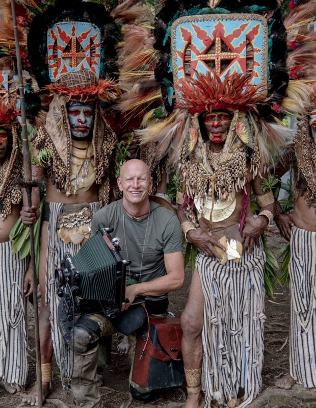 A person crouches, holding a camera, surrounded by individuals wearing traditional, colorful feathered headdresses and ornate costumes. The background features a natural setting with vegetation.