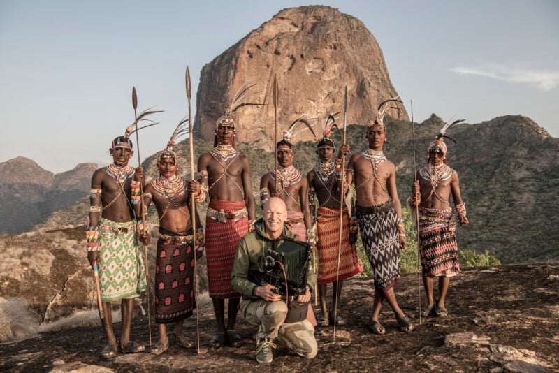 A group of traditionally dressed individuals stand with a photographer in front of a rocky mountain landscape. They hold spears and wear colorful attire, adorned with beads and headpieces. The photographer kneels in front, holding a camera.