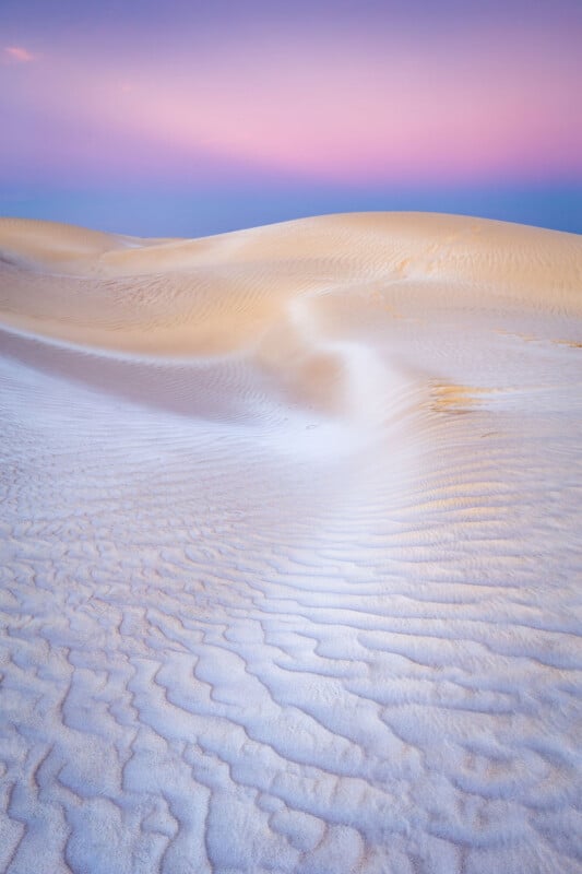 A desert landscape with rippled sand dunes under a purple and pink sky. The smooth curves and gentle shadows create a serene and tranquil atmosphere.