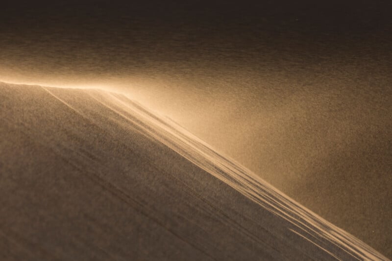 Wind-blown sand creates a soft, golden wave pattern over a dune. The light and shadow highlight the gentle curves and texture, evoking a serene and ethereal desert landscape.