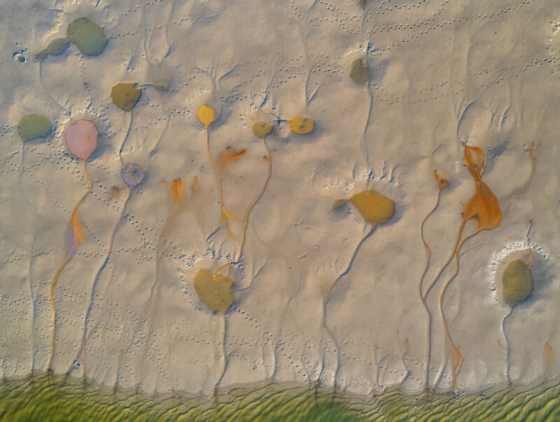 Aerial view of a sandy beach with abstract shapes and patterns. Various small puddles create organic, colorful forms resembling paint splatters. Wavy lines at the bottom suggest tidal influences. Subtle footprints can be seen across the sand.