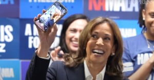 A woman in a suit holds up a smartphone displaying a selfie of herself with a beaming smile. She is surrounded by people in a room decorated with blue campaign signs.