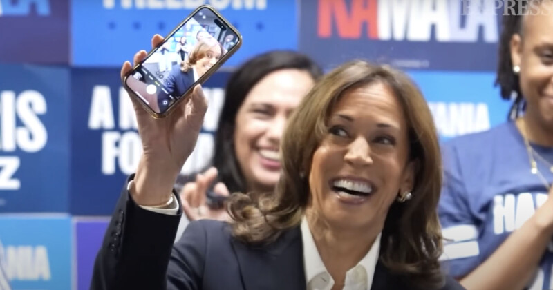 A woman in a suit holds up a smartphone displaying a selfie of herself with a beaming smile. She is surrounded by people in a room decorated with blue campaign signs.