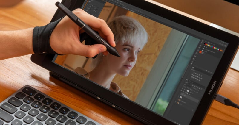 A person uses a stylus to edit a portrait photo of a woman on a tablet with photo editing software. A keyboard is visible on the wooden desk.