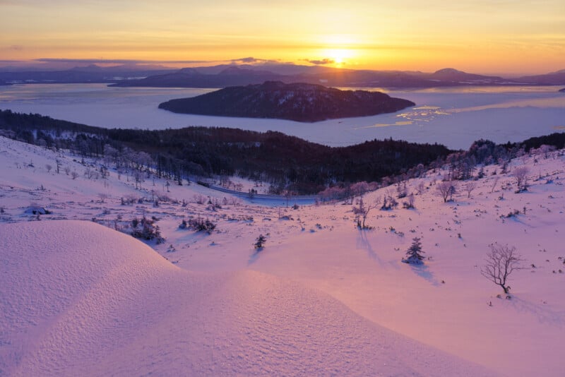 A stunning winter sunrise over a snow-covered landscape with rolling hills in the foreground. A distant frozen lake and mountains are silhouetted against the vibrant orange and purple sky. Sparse trees dot the serene scene.