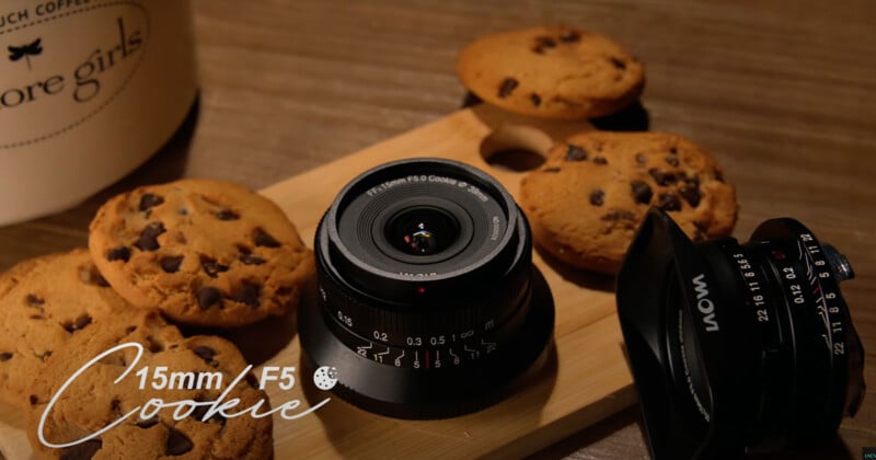 A camera lens is displayed on a wooden board surrounded by chocolate chip cookies. A camera lens cap is nearby. Text on the image reads "15mm F/5 Cookie." A jar labeled "more girls" is partially visible in the background.