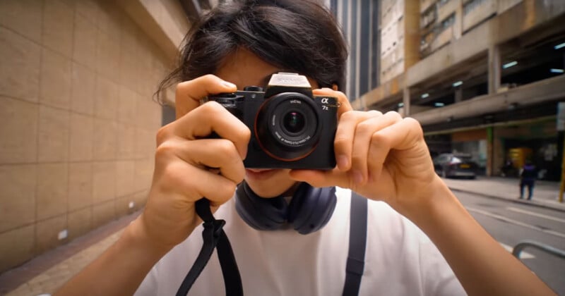 A person taking a photo with a camera on a city street. They have headphones around their neck and are wearing a white shirt. Urban buildings and a multi-story car park are visible in the background.