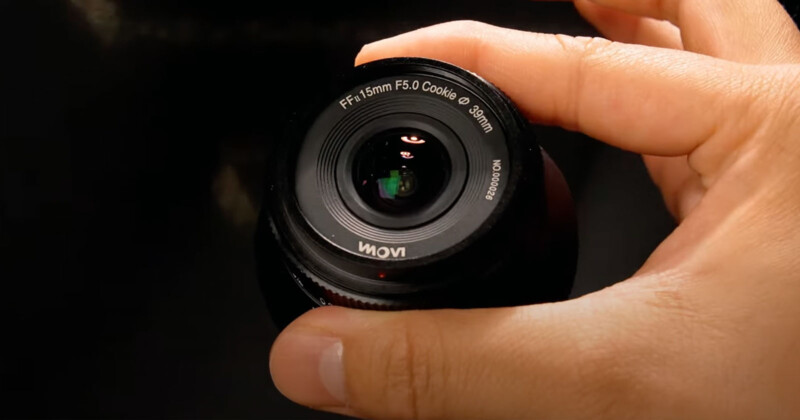 A hand holding a black camera lens labeled "FF-15mm F5.0 Cookie" against a dark background. The lens has reflective glass and a visible red alignment dot. The hand is positioned to show the front view of the lens.