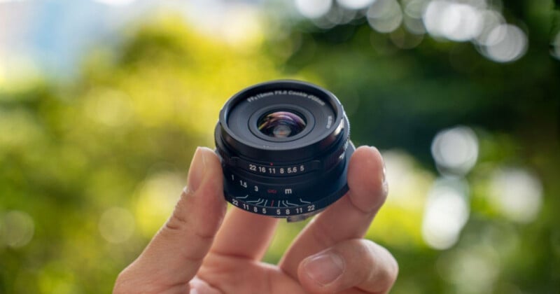 A hand is holding a compact camera lens against a blurred outdoor background with greenery. The lens details and focus rings are visible.