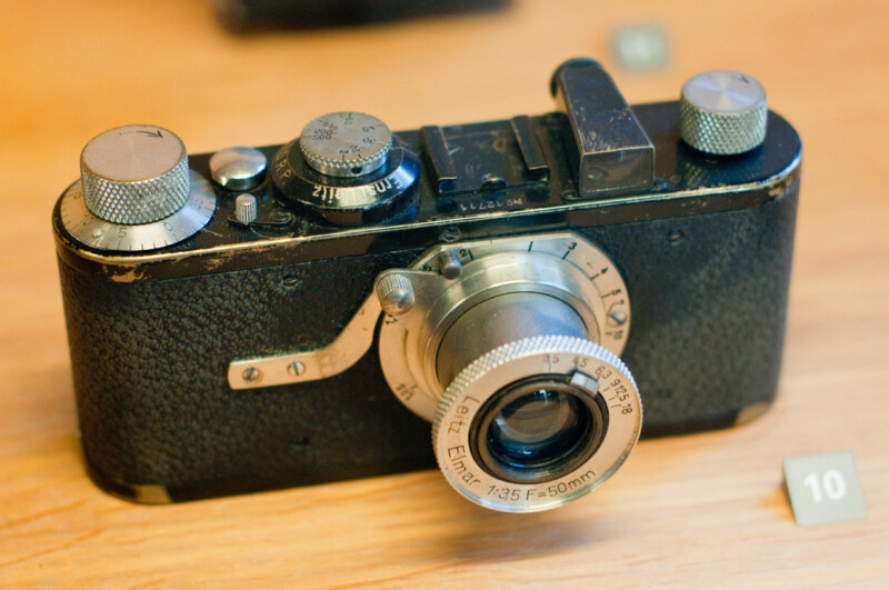 A vintage black Leica camera with a silver lens and dials, displayed on a wooden surface. The camera's metal parts show signs of wear, and there is a small numbered tag beside it.