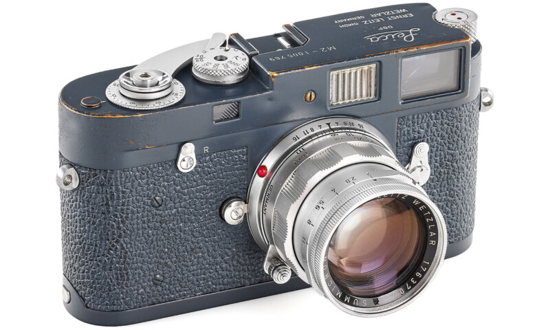 A vintage Leica film camera with a textured black body and metallic details. The lens is prominently featured, displaying various markings. The camera shows signs of wear, highlighting its age and use.