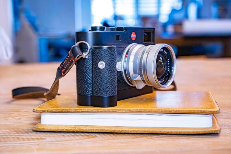 A vintage-style camera with a leather strap rests on a closed, tan notebook on a wooden table. The setting appears to be indoors with soft, diffused lighting.