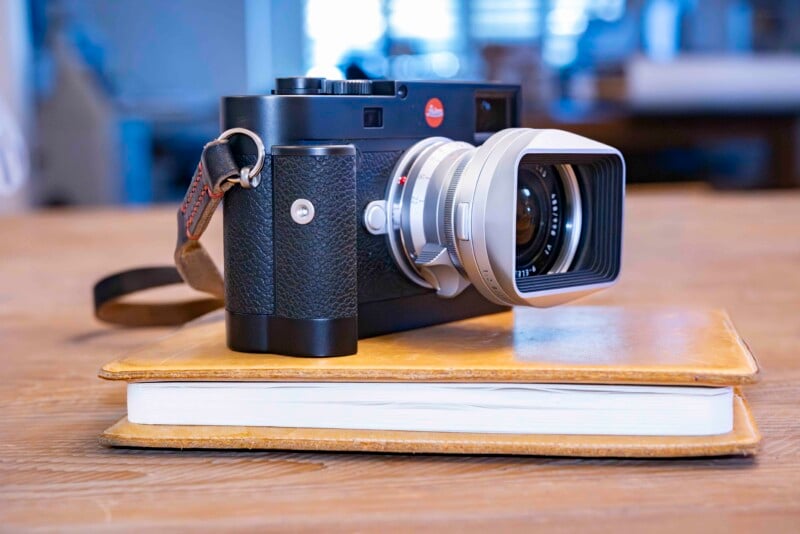 A black and silver vintage-style camera rests on top of a closed, yellow notebook on a wooden table. The camera has a textured grip and a wide lens with a rectangular hood. The setting is softly lit, suggesting an indoor environment.