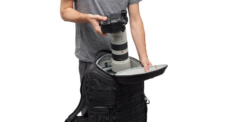 A person in a gray shirt is placing a large camera with a telephoto lens into a black camera backpack. The bag is open, revealing compartments for equipment. The person is partially visible, emphasizing the focus on the camera gear.