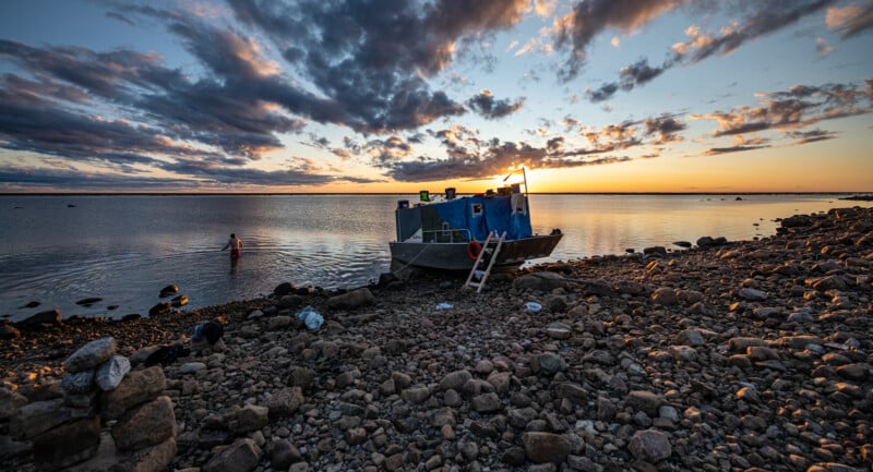 A person standing in the water near a rocky shoreline at sunset. A small, rustic boat is on the shore with a ladder leaning against it. The sky is colorful with clouds reflecting the setting sun over the calm ocean.