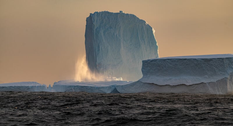 A large iceberg stands majestically in the ocean at sunset. A misty spray is visible around its base, and the warm light casts a golden glow on the ice. The foreground shows dark, rippling water.