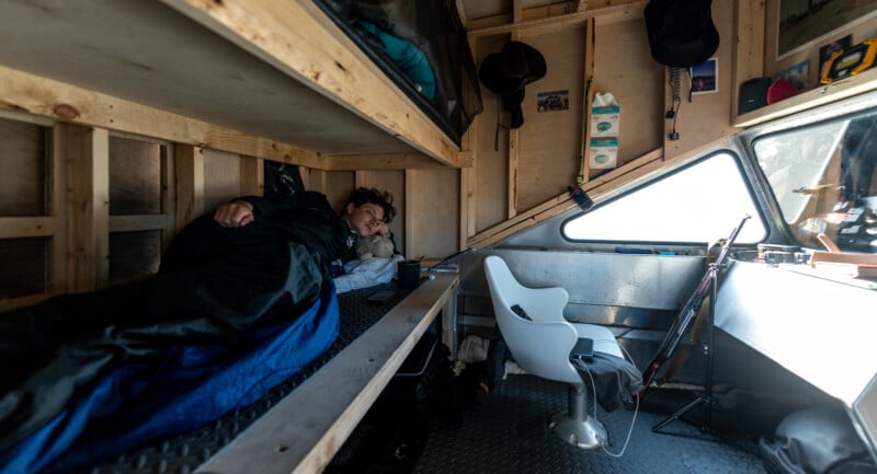 A person sleeping in a cozy, wooden interior of a small boat. The space features a bunk bed, various personal items, a guitar, a skateboard, and a white chair near the window. Sunlight streams in through the window, illuminating the area.