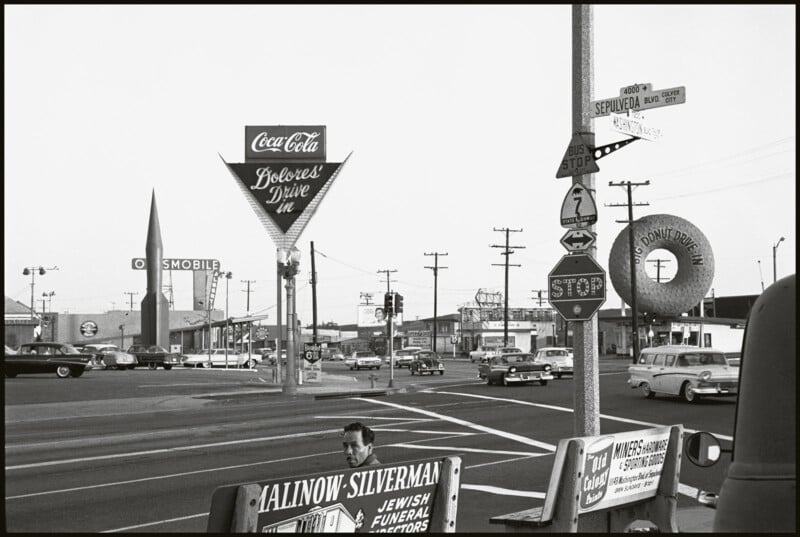 A vintage 1960s street scene with classic cars at an intersection. Signs for Coca-Cola, Silver's Drive In, and various street directions are visible. An Oldsmobile dealership and Goodyear tire sign are seen in the background.