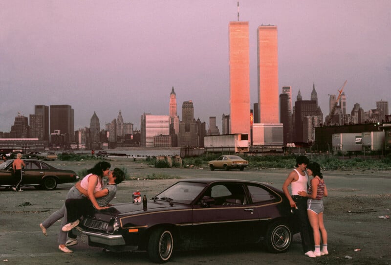 A couple sits on the hood of a car, affectionately embracing, with two onlookers nearby. The skyline of downtown Manhattan, including the Twin Towers, looms in the background under a pink sky. A soda bottle rests on the car's hood.