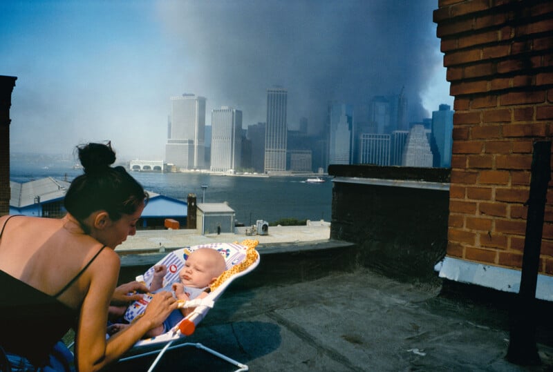 A woman tends to a baby on a rooftop while smoke rises from the New York City skyline in the background. The scene captures a juxtaposition of nurturing care and a monumental event.