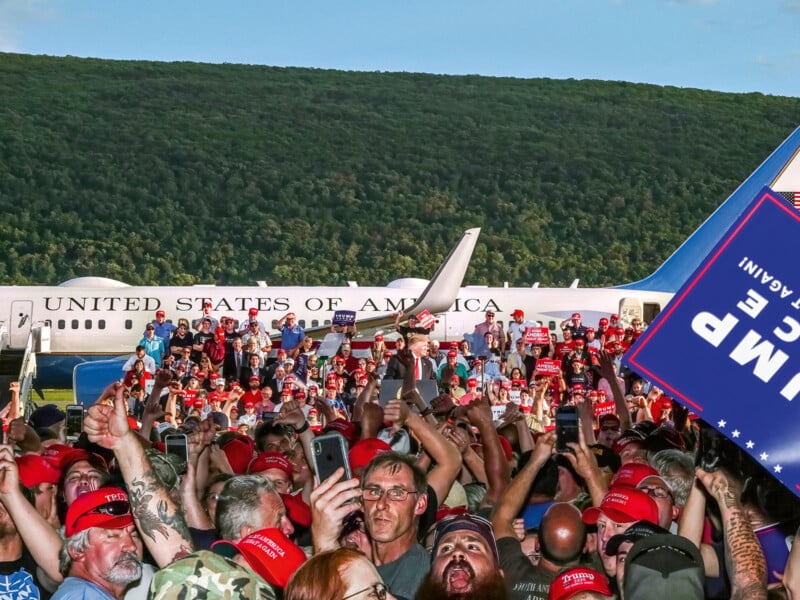 A large crowd wearing red hats and holding signs gathers near an airplane with "United States of America" written on it. A forested hill is visible in the background. People are taking photos with their phones.