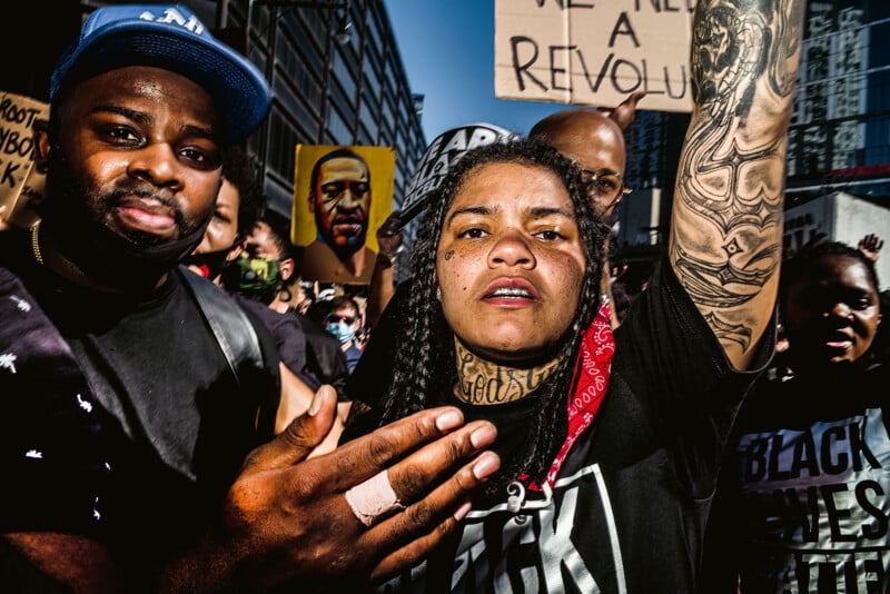 A diverse group of protesters in an urban setting, holding signs advocating for racial justice. The focus is on a person with tattoos and a red bandana, gesturing towards the camera. The crowd shows unity and determination.