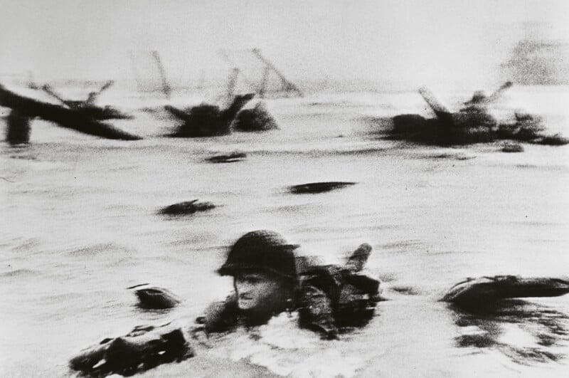 A blurred black-and-white photo shows a soldier wading through water, surrounded by debris and the silhouette of landing crafts in the background. The scene conveys movement and urgency, possibly set during a historic amphibious assault.