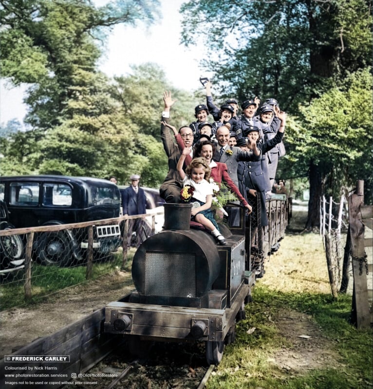 A group of people, including a child holding a bouquet, ride on a small open train outdoors. They are smiling and waving, surrounded by trees and parked cars. The scene feels joyful and celebratory.