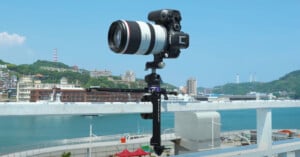 A professional camera with a large lens mounted on a tripod stands on a rooftop railing, overlooking a scenic view of a harbor with boats, buildings, and hills under a clear blue sky.