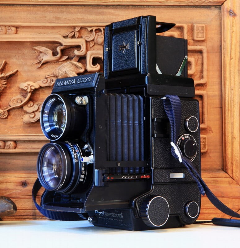 A vintage Mamiya C330 twin-lens reflex camera with a black body and textured grips is displayed against a carved wooden backdrop. The camera features two lenses aligned vertically and a shoulder strap.