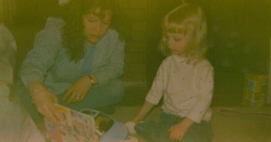 A woman and a young girl sit on the floor. The woman is holding a colorful book, showing it to the girl. The room is dimly lit, and there's a stack of colorful blocks in the background.