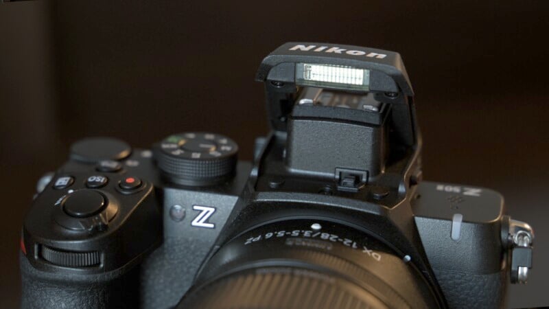 A close-up of a Nikon Z series camera shows the camera's top controls and an open flash unit. The lens is partially visible, and the focus is on the camera's buttons and flash mechanism. The background is blurred and dark.