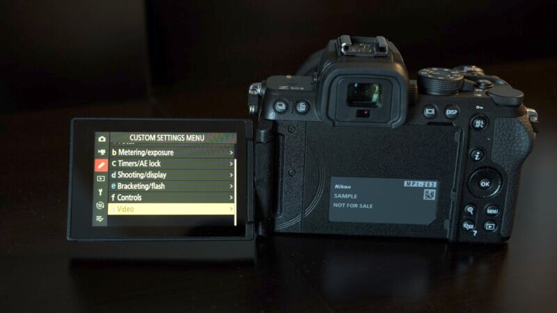 A digital camera with a flip-out screen showing the custom settings menu. Options include metering exposure, timers, shooting display, bracketing/flash, controls, and video. The screen displays a sample message and a model identifier.