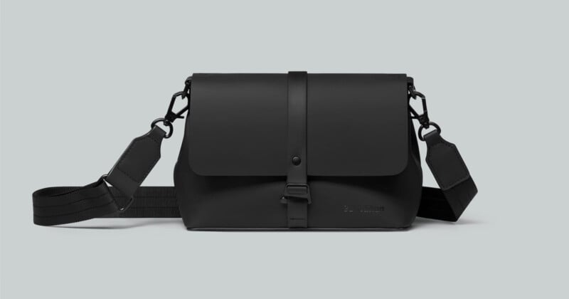 A sleek black crossbody bag with a flap closure and adjustable strap is centered against a light gray background. The bag has minimalistic design elements, including a subtle embossed logo on the front.