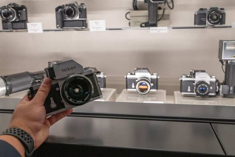 A person holds a classic Nikon camera in front of a display case featuring various vintage cameras. The case includes different models and a large lens, all showcased under soft lighting.