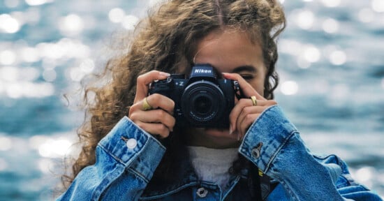 A person with curly hair and wearing a denim jacket holds a Nikon camera up to their face, capturing a photo. The background is blurred with sparkling water.