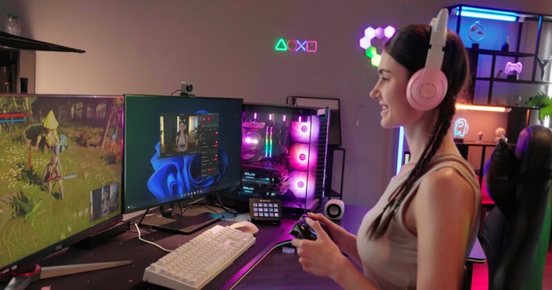 A person wearing pink headphones is sitting at a desk, playing a video game on a dual-monitor setup. They hold a gaming controller and colorful LED lights illuminate the room. Various gaming accessories and decor are visible in the background.