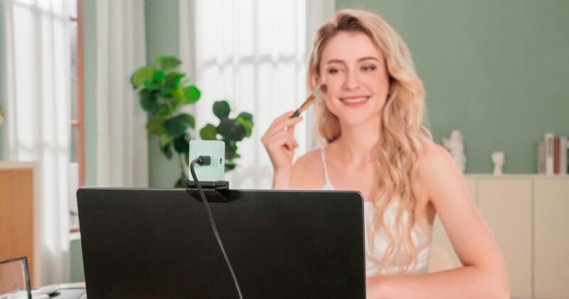 A person with long blonde hair is smiling while applying makeup in front of a laptop with a webcam. The room is softly lit, with a plant and some shelves in the background.