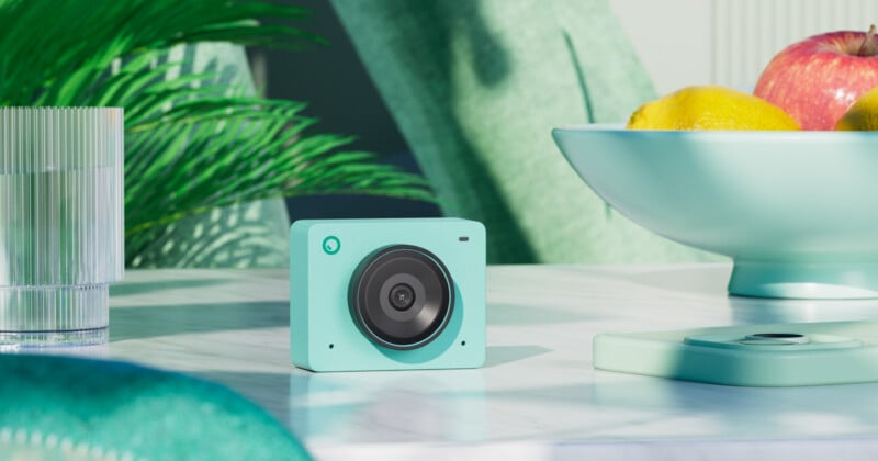 A small, modern teal camera sits on a white table next to a matching teal lid. A glass of water, a bowl of apples and lemons, and green foliage in the background create a fresh, vibrant setting.