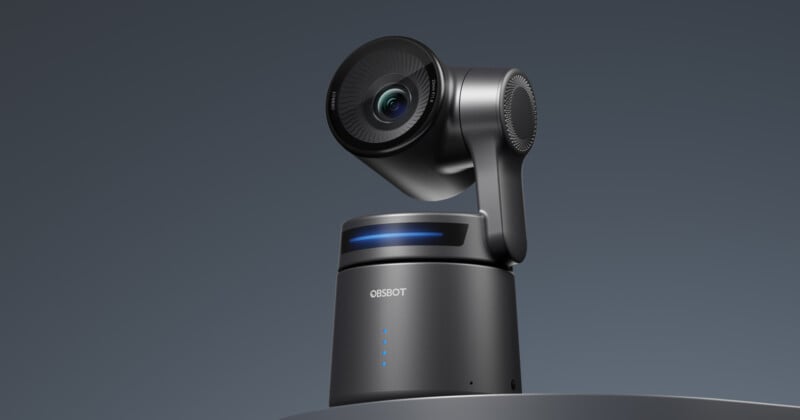 A sleek black robotic camera with a cylindrical base and rotating lens is set against a dark background. The base features a blue illuminated strip and LED indicators.