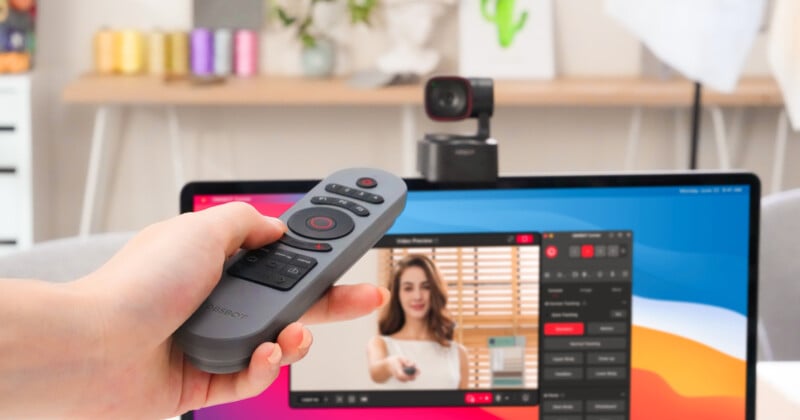 A person is holding a remote control in front of a computer monitor with a webcam mounted on top. The screen displays a woman adjusting a curtain, using software for video recording. The room has a soft, bright interior with decorative elements.